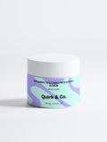 A white cosmetic jar of Quirk & Co. Face & Body Scrub with a colorful label featuring purple and mint green abstract waves, set against a clean white background.